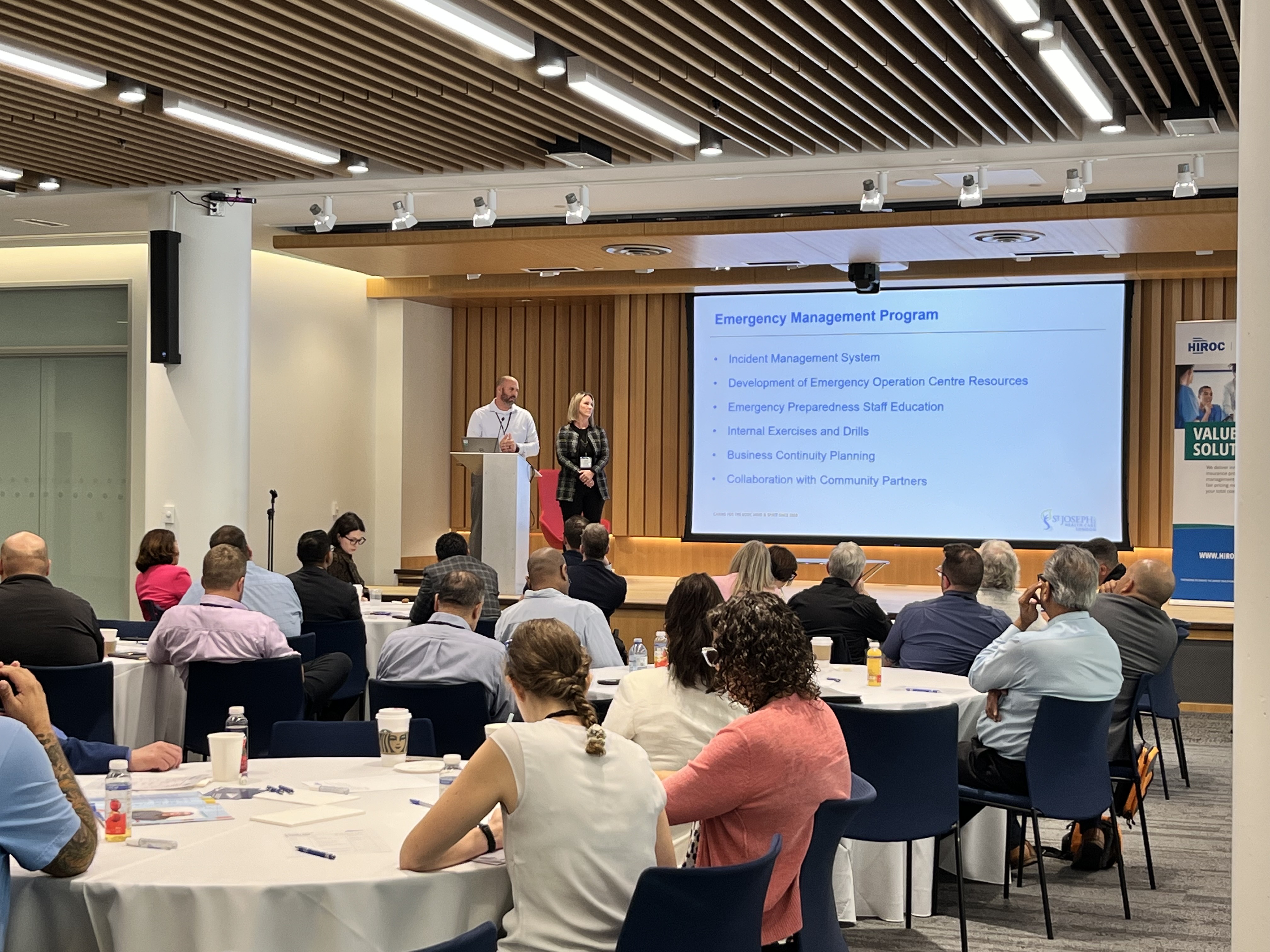 Presenters Peter Kreis, left, and Danica Low from St. Joseph’s Health Care London stand on stage at the podium and speak to a full room of attendees.