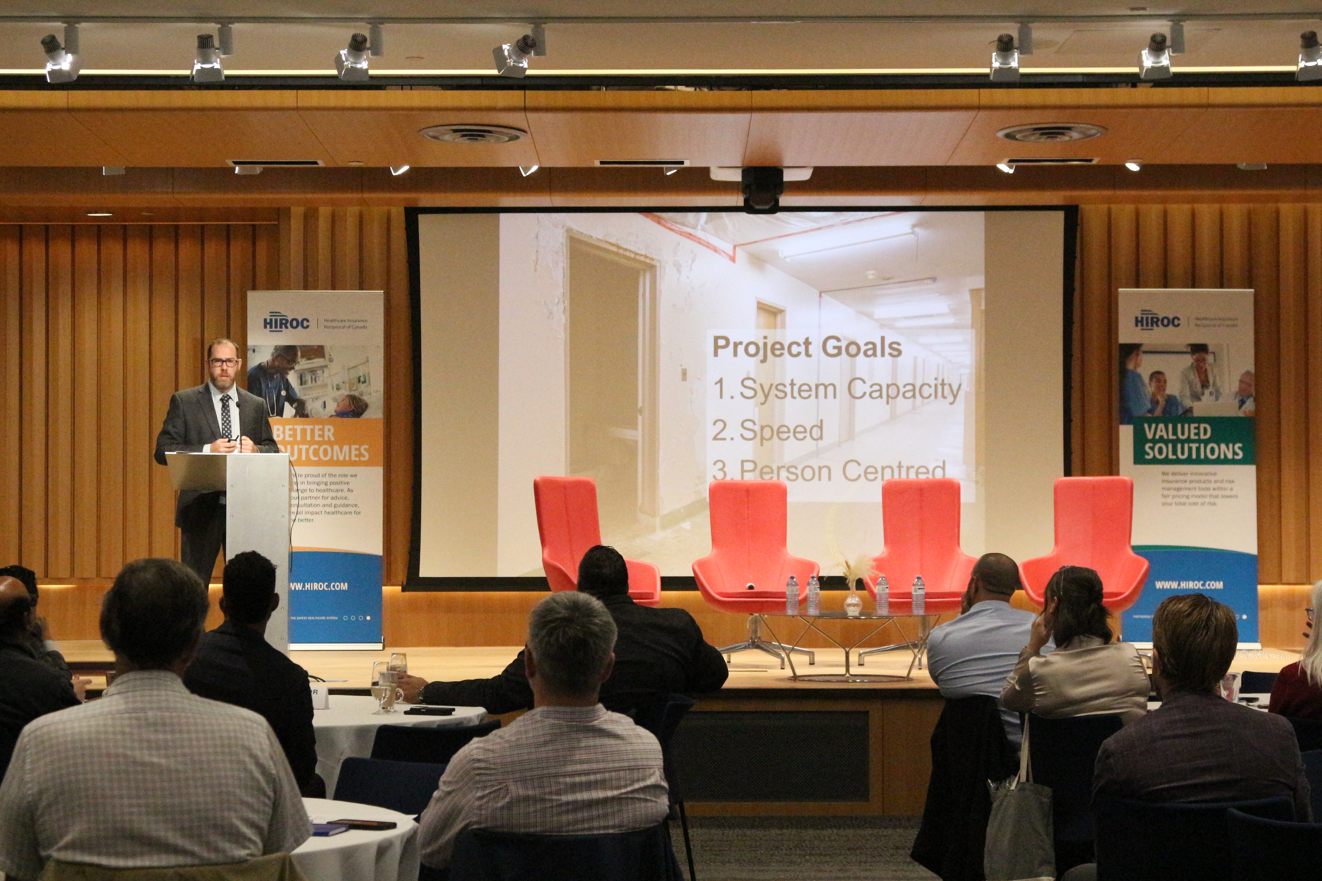 Speaker on stage next to large screen and audience in front