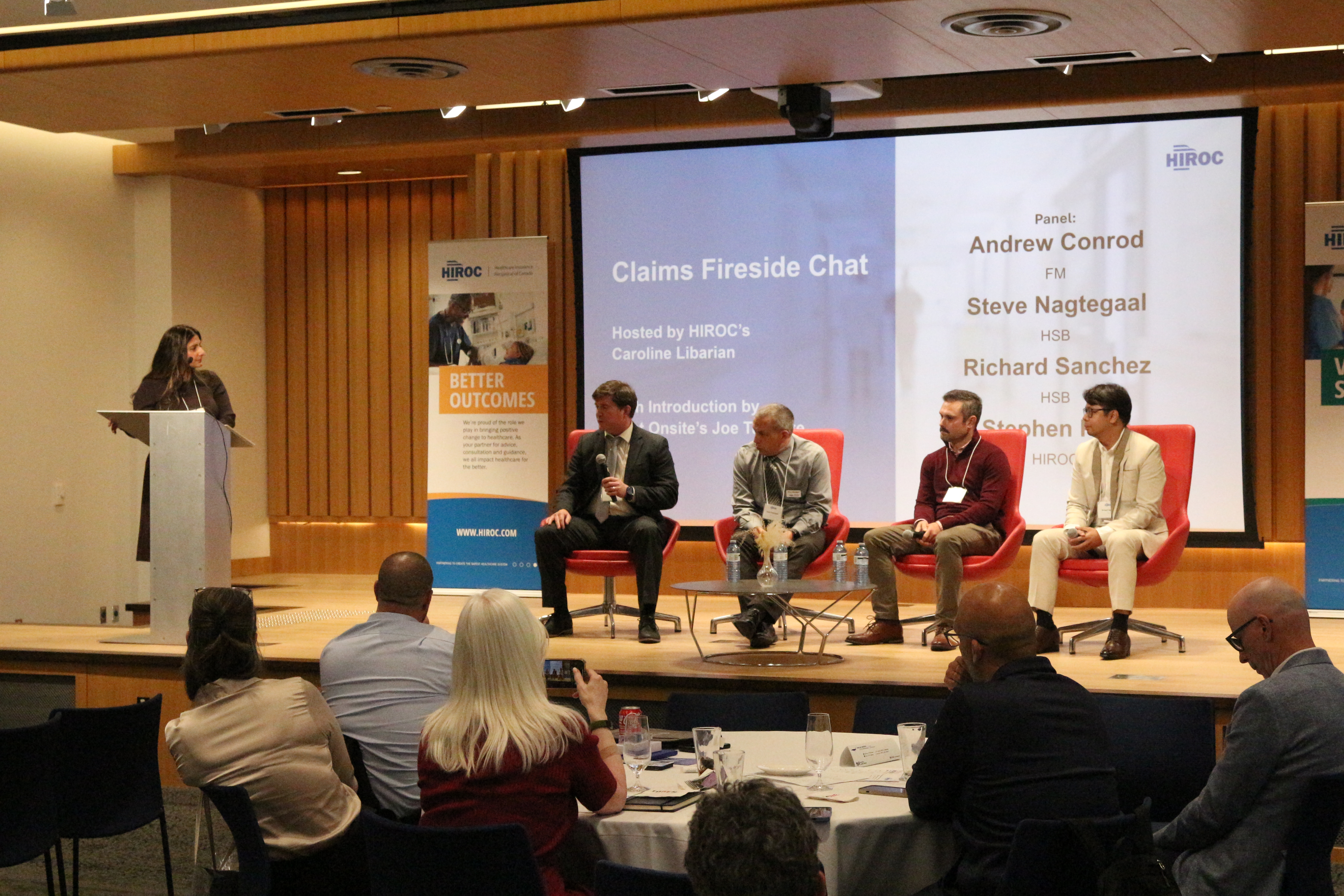 Panel speakers sit on stage in front of a large screen while moderator stands at podium