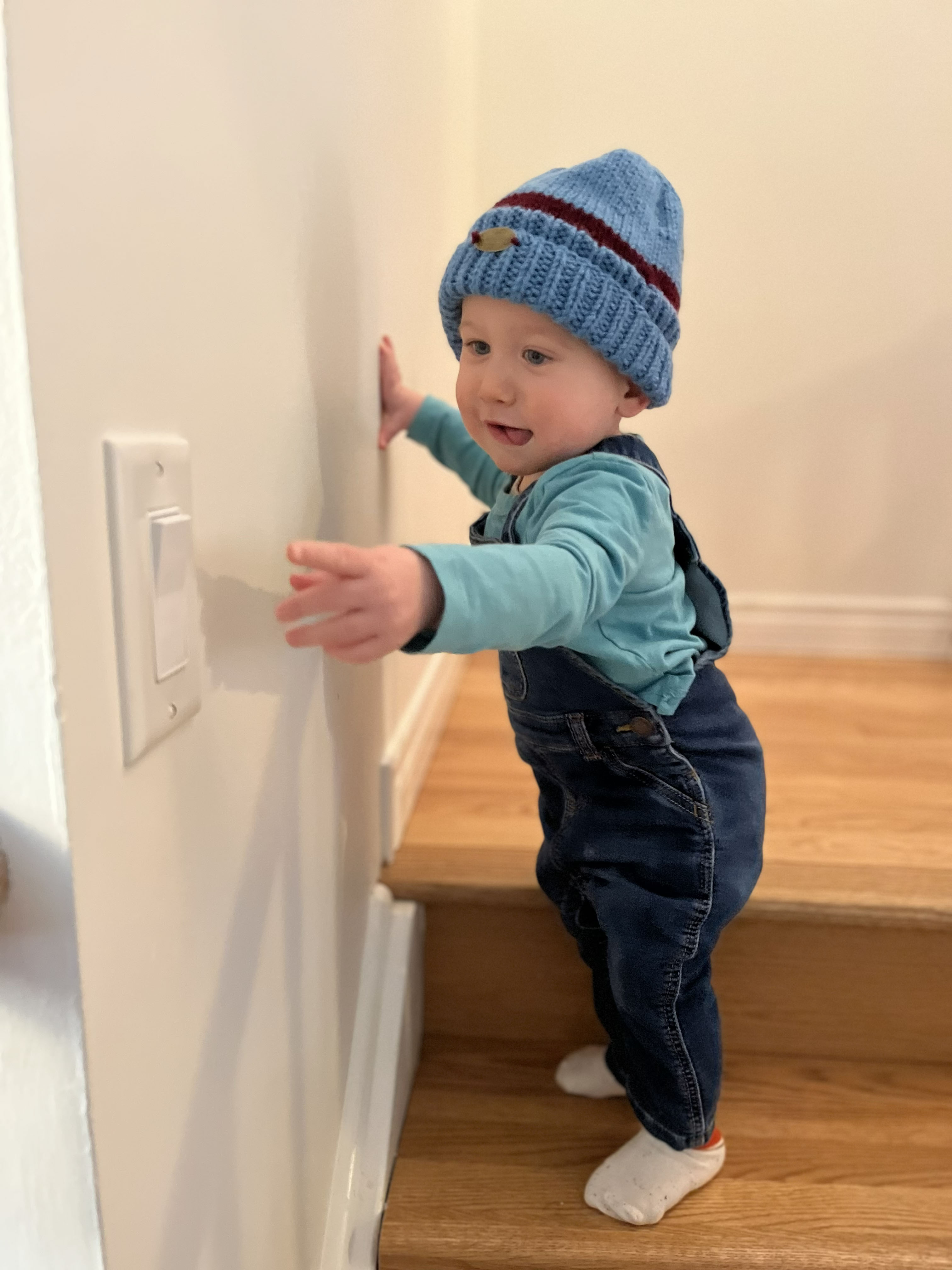 A toddler smiling and leaning against a wall while wearing a blue knit hat made by Catherine.