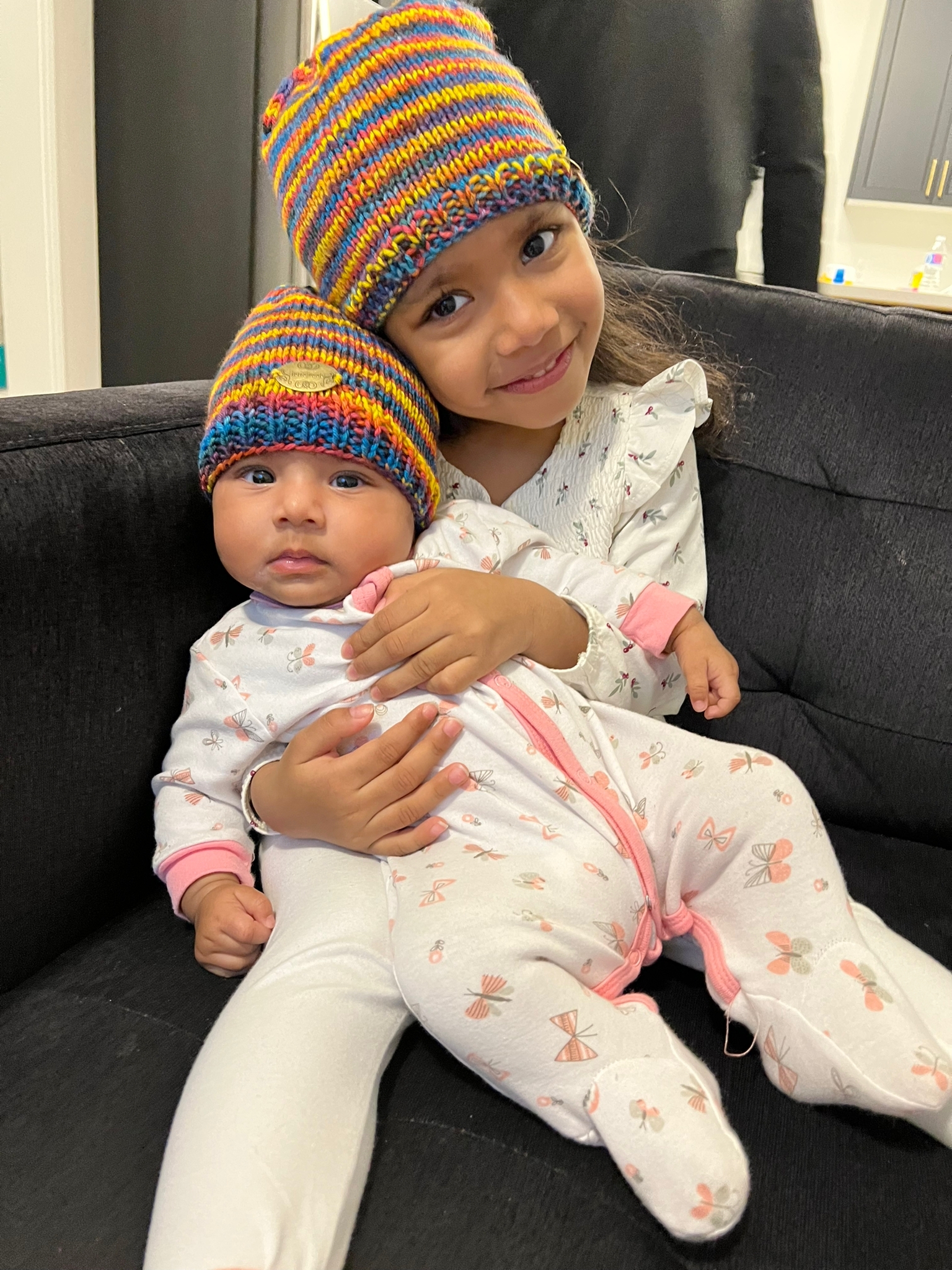 A little kid hugging her baby sibling and both are wearing Catherine's multicoloured, striped knit hats.