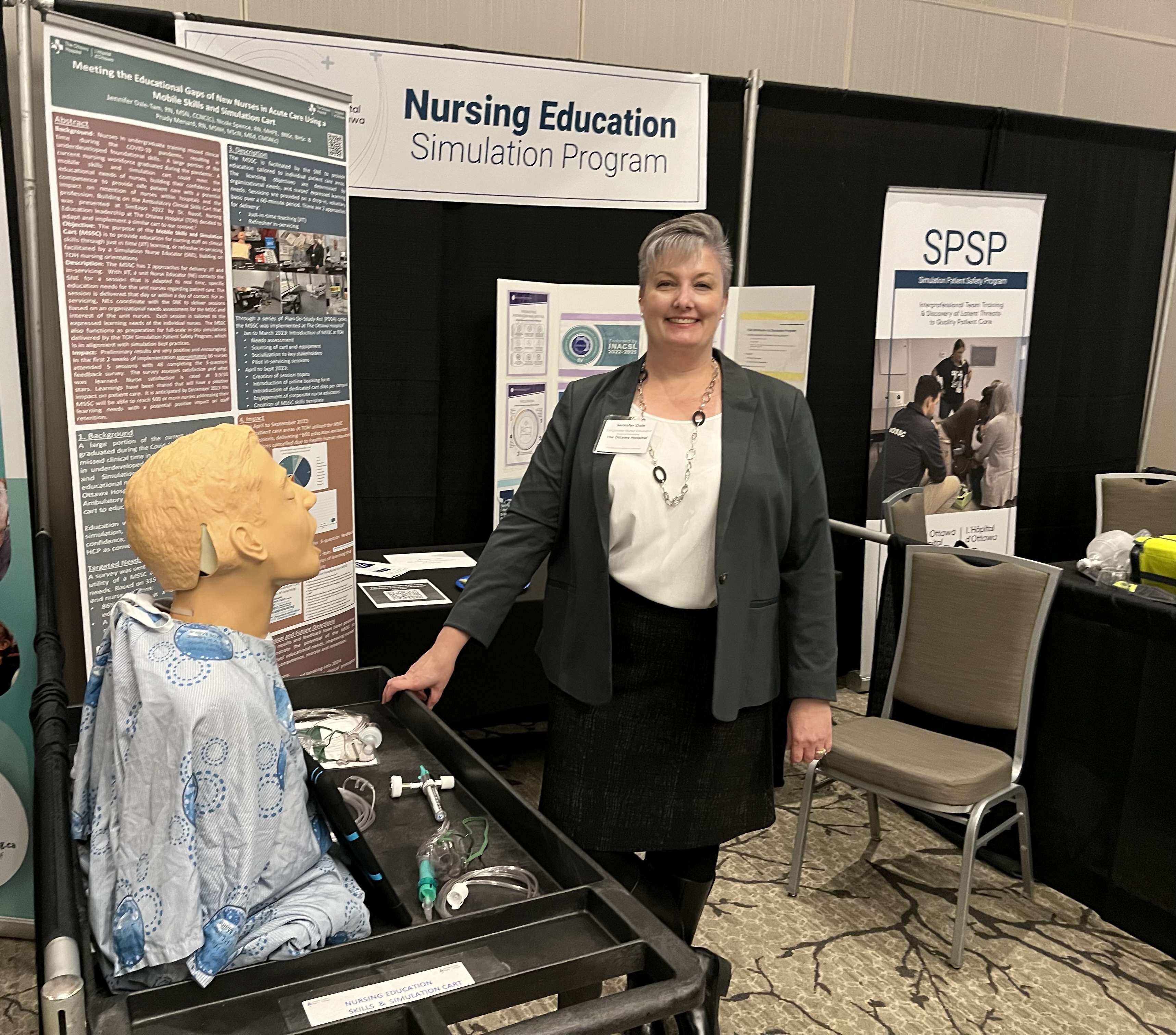 Jennifer Dale-Tam smiling in her booth for TOH's Simulation Patient Safety Program at a conference with program banners behind her..