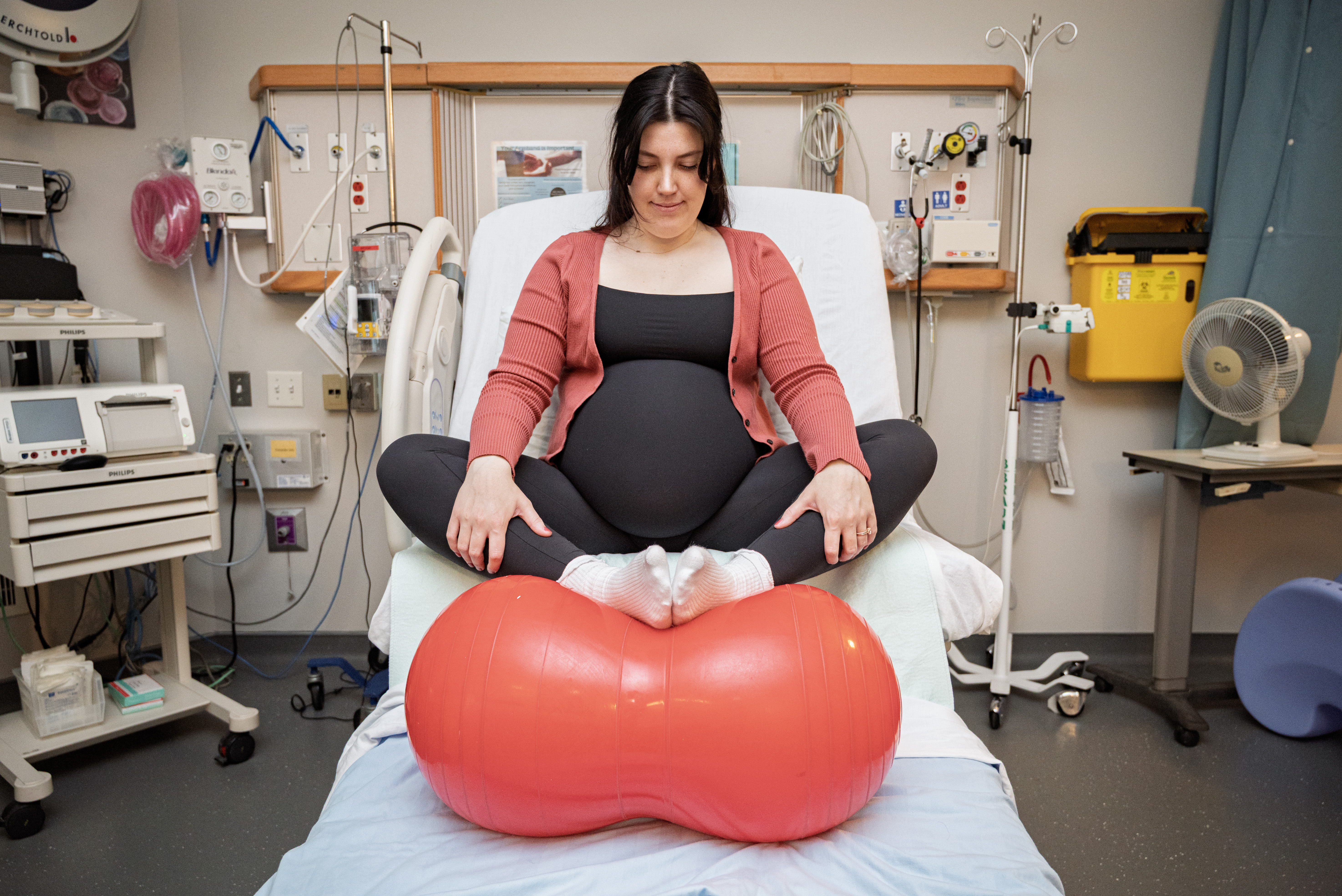 A pregnant person sits on a hospital bed, feet up on a medicine ball.