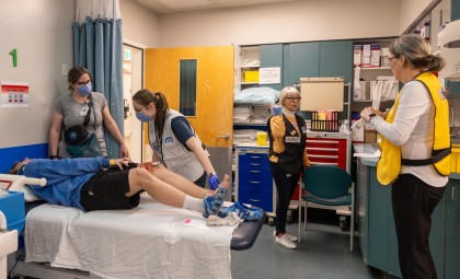 Simulated patient lies on bed and several healthcare workers are in the room